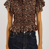 A woman wearing a brown leopard-print smocked blouse with ruffled sleeves and a ruffled hem, paired with dark blue jeans, against a plain light background.