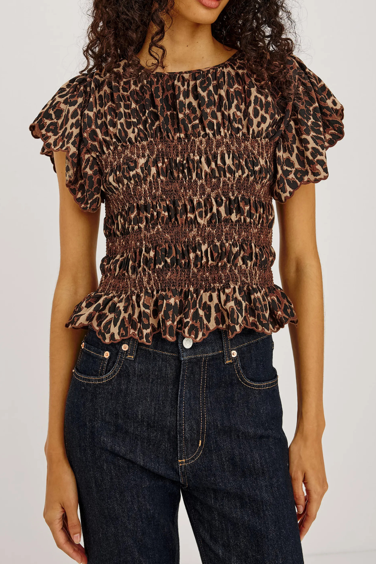 A woman wearing a brown leopard-print smocked blouse with ruffled sleeves and a ruffled hem, paired with dark blue jeans, against a plain light background.