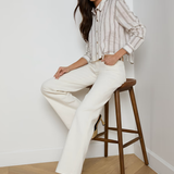 Woman sitting on a wooden stool wearing a striped shirt and white pants in a minimalistic setting.