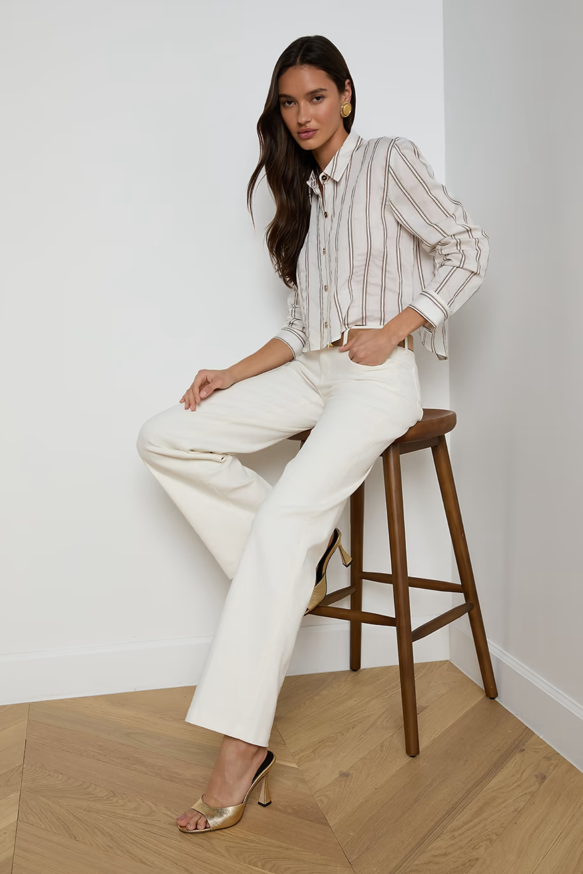 Woman sitting on a wooden stool wearing a striped shirt and white pants in a minimalistic setting.