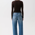 A woman viewed from behind wearing a dark brown long-sleeve top, blue wide-leg jeans, and dark brown pointed-toe heels against a plain light gray studio background.