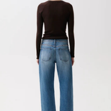 A woman viewed from behind wearing a dark brown long-sleeve top, blue wide-leg jeans, and dark brown pointed-toe heels against a plain light gray studio background.
