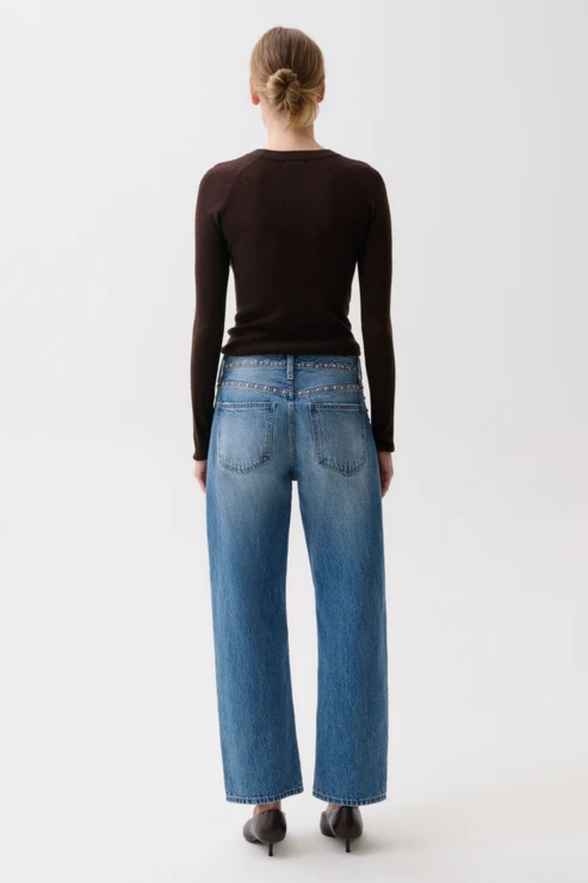 A woman viewed from behind wearing a dark brown long-sleeve top, blue wide-leg jeans, and dark brown pointed-toe heels against a plain light gray studio background.