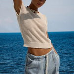 Woman in a beige top and light blue shorts standing by the ocean.
