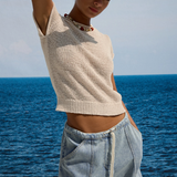 Woman in a beige top and light blue shorts standing by the ocean.