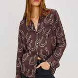 A woman modeling a brown button-up blouse with a white horseshoe pattern, paired with dark jeans, against a light gray studio background.