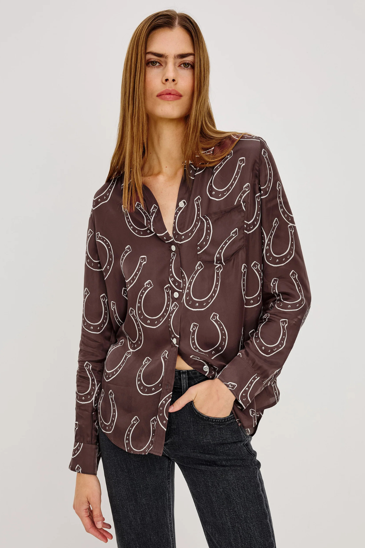 A woman modeling a brown button-up blouse with a white horseshoe pattern, paired with dark jeans, against a light gray studio background.