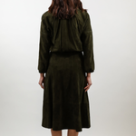 A woman viewed from behind wearing an olive green two-piece set consisting of a cropped long-sleeve top and a matching midi skirt, paired with white heels against a plain light gray studio background.