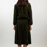 A woman viewed from behind wearing an olive green two-piece set consisting of a cropped long-sleeve top and a matching midi skirt, paired with white heels against a plain light gray studio background.