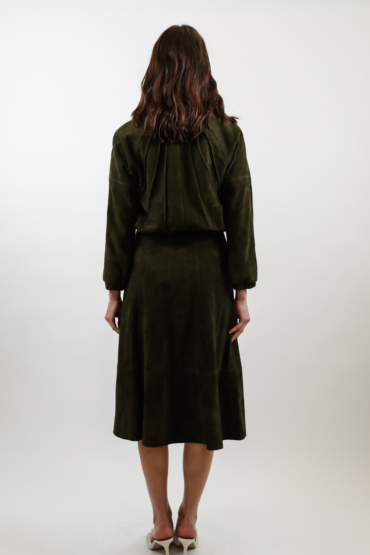 A woman viewed from behind wearing an olive green two-piece set consisting of a cropped long-sleeve top and a matching midi skirt, paired with white heels against a plain light gray studio background.