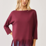 A woman wearing a boat-neck, three-quarter sleeve top in deep burgundy with fringe trim at the hem, paired with light blue jeans, against a plain light gray background.