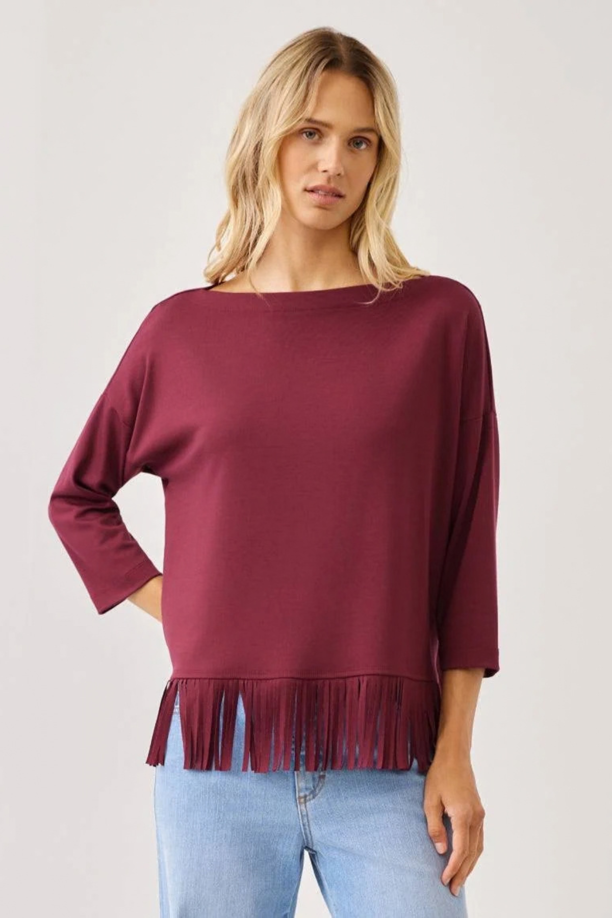 A woman wearing a boat-neck, three-quarter sleeve top in deep burgundy with fringe trim at the hem, paired with light blue jeans, against a plain light gray background.