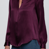 A woman wearing a long-sleeve satin blouse in deep plum purple with a split collar and button front, paired with blue jeans, against a light gray studio background.