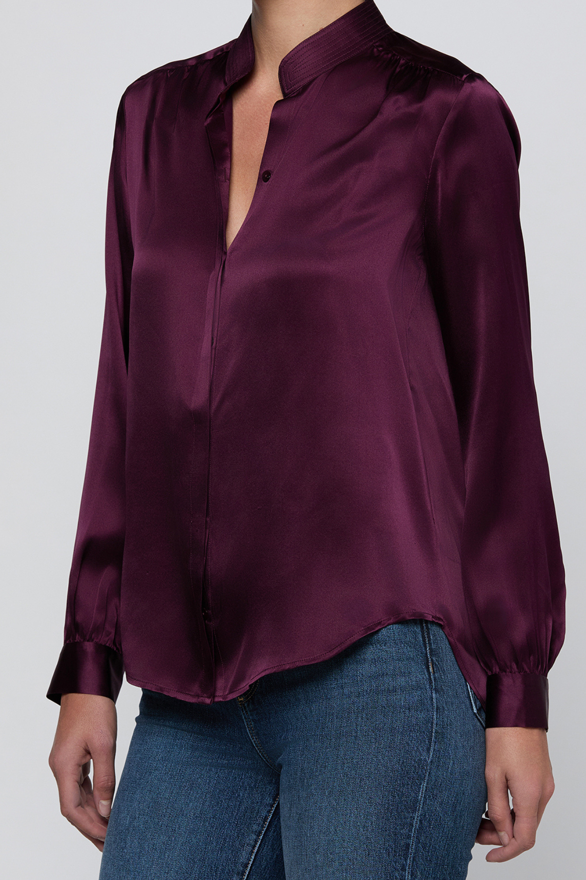 A woman wearing a long-sleeve satin blouse in deep plum purple with a split collar and button front, paired with blue jeans, against a light gray studio background.