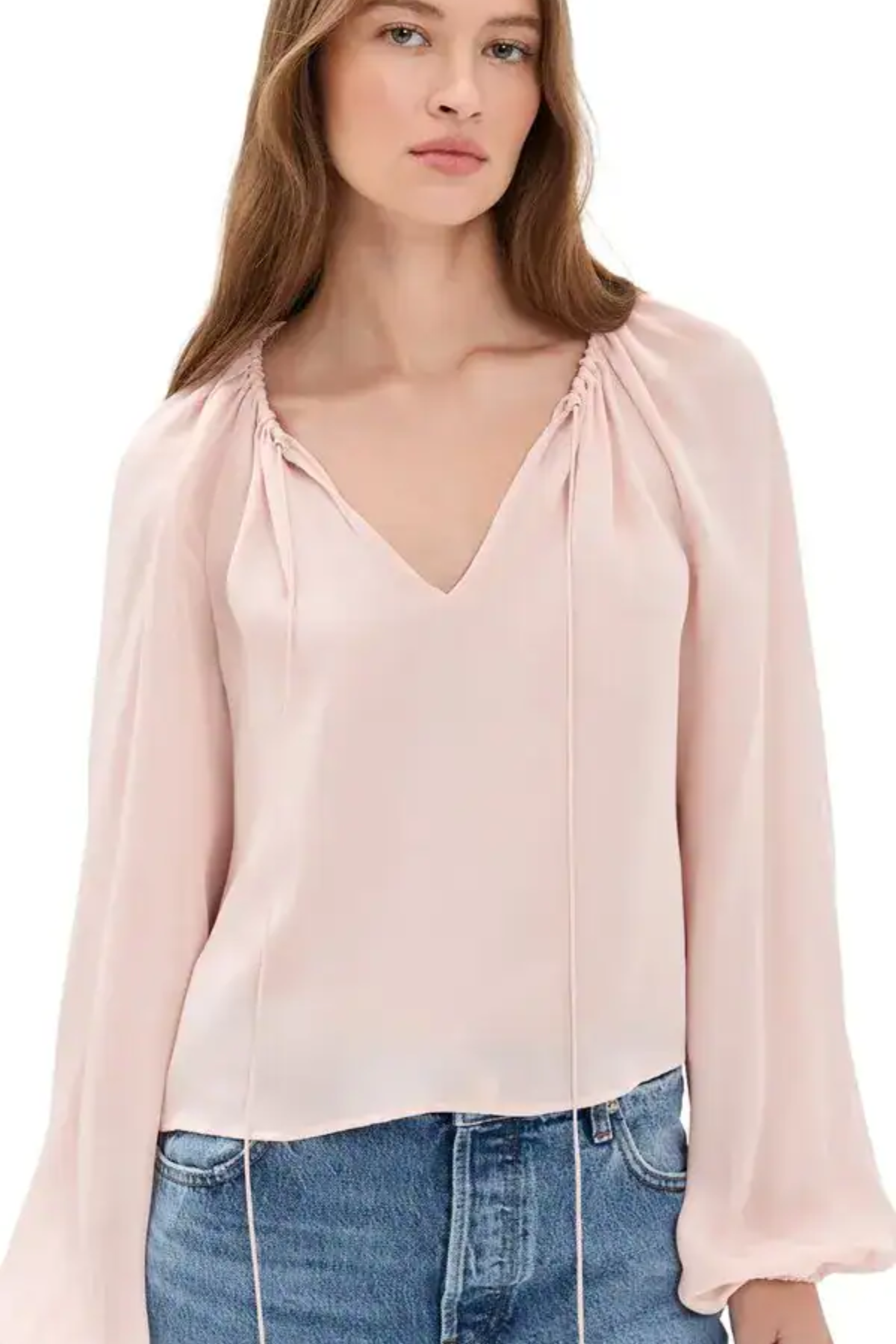 A woman wearing a light pink long-sleeve blouse with a gathered V-neckline and drawstring details, paired with blue jeans, against a plain white background.