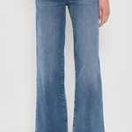 A person wearing medium-wash blue wide-leg flare jeans with a faded wash, paired with beige woven flat shoes, shown from the waist down against a plain white studio background.