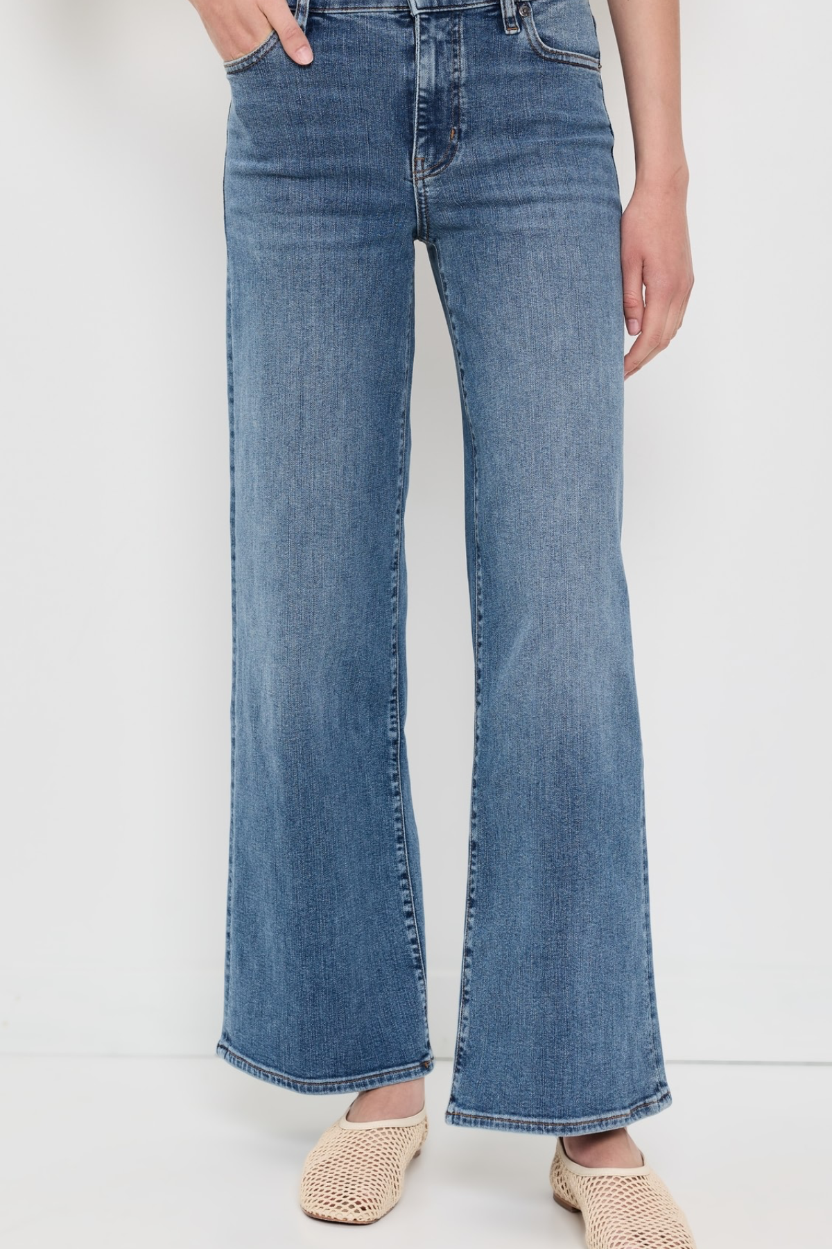 A person wearing medium-wash blue wide-leg flare jeans with a faded wash, paired with beige woven flat shoes, shown from the waist down against a plain white studio background.