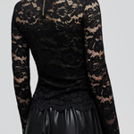 A woman shown from the back wearing a black long-sleeve lace top with a high mock neck and a small gold zipper at the collar, paired with black pants, against a light gray studio background.