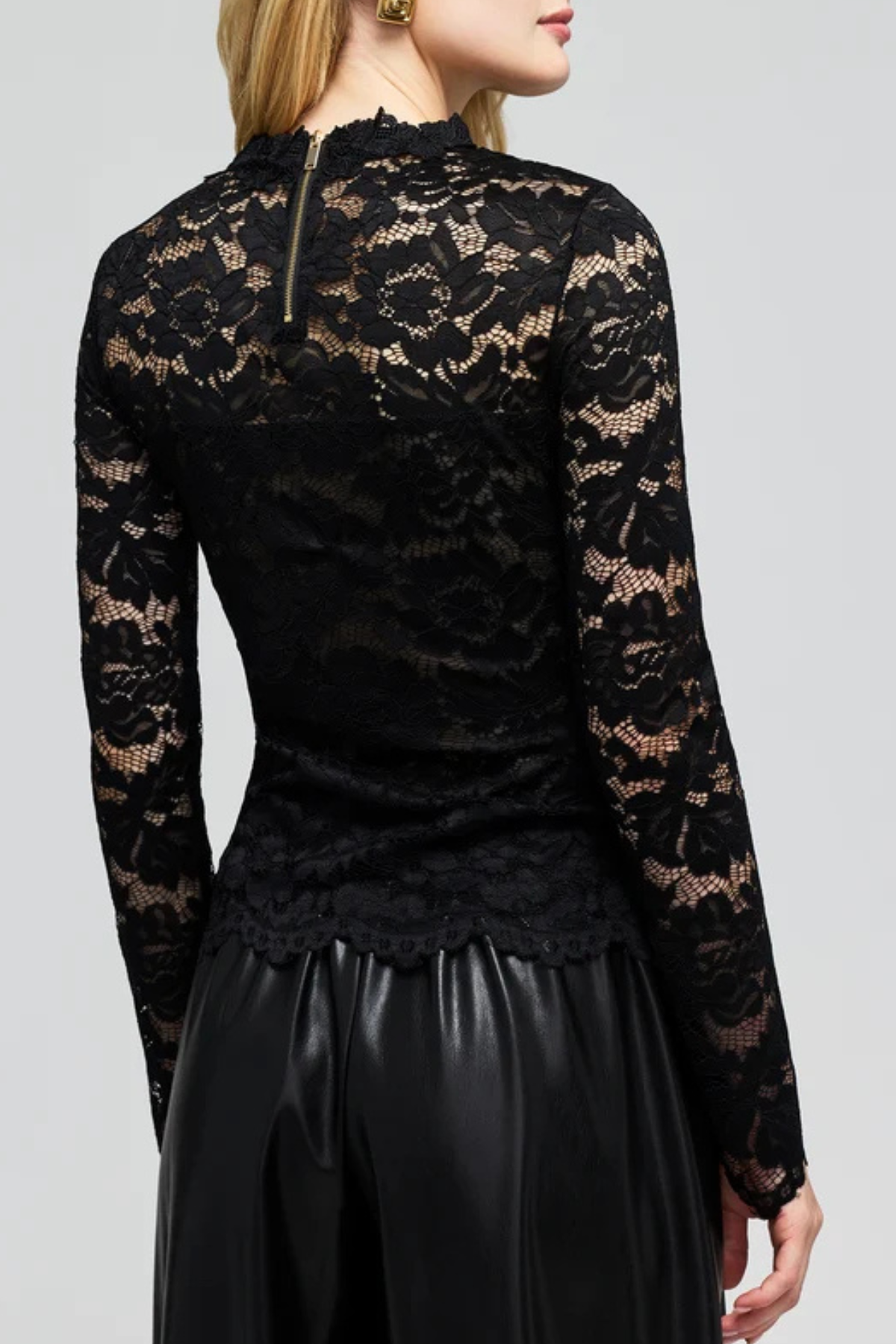 A woman shown from the back wearing a black long-sleeve lace top with a high mock neck and a small gold zipper at the collar, paired with black pants, against a light gray studio background.