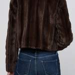 A person shown from the back wearing a dark brown cropped fur jacket with vertical stitched paneling and a plush collar, paired with dark blue jeans, against a light gray studio background.