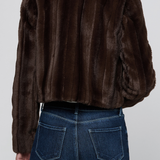A person shown from the back wearing a dark brown cropped fur jacket with vertical stitched paneling and a plush collar, paired with dark blue jeans, against a light gray studio background.