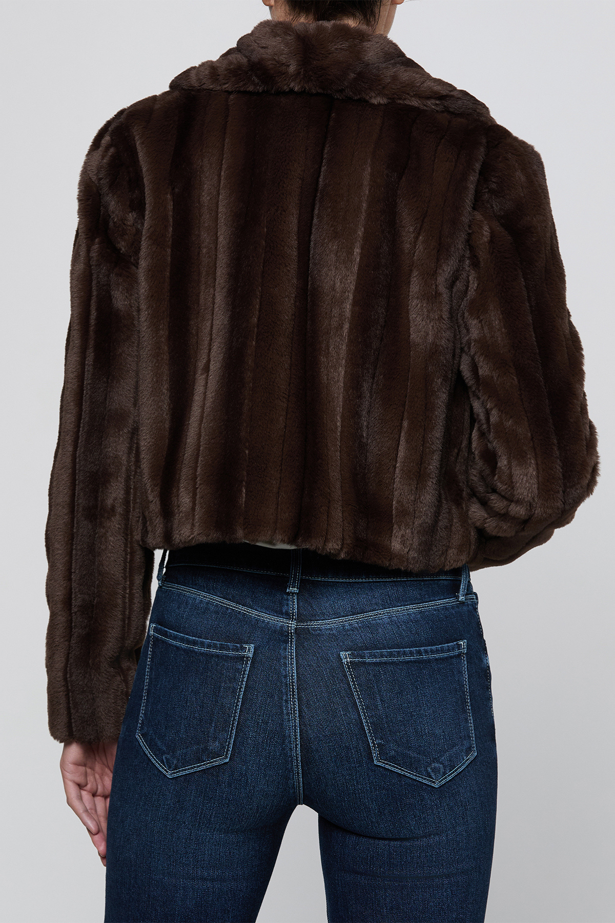 A person shown from the back wearing a dark brown cropped fur jacket with vertical stitched paneling and a plush collar, paired with dark blue jeans, against a light gray studio background.