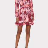 A model wearing a long-sleeve wrap-style mini dress with a pink, red, and gray floral pattern, featuring a tie waist and ruffled hem, paired with clear high-heeled sandals against a light gray background.