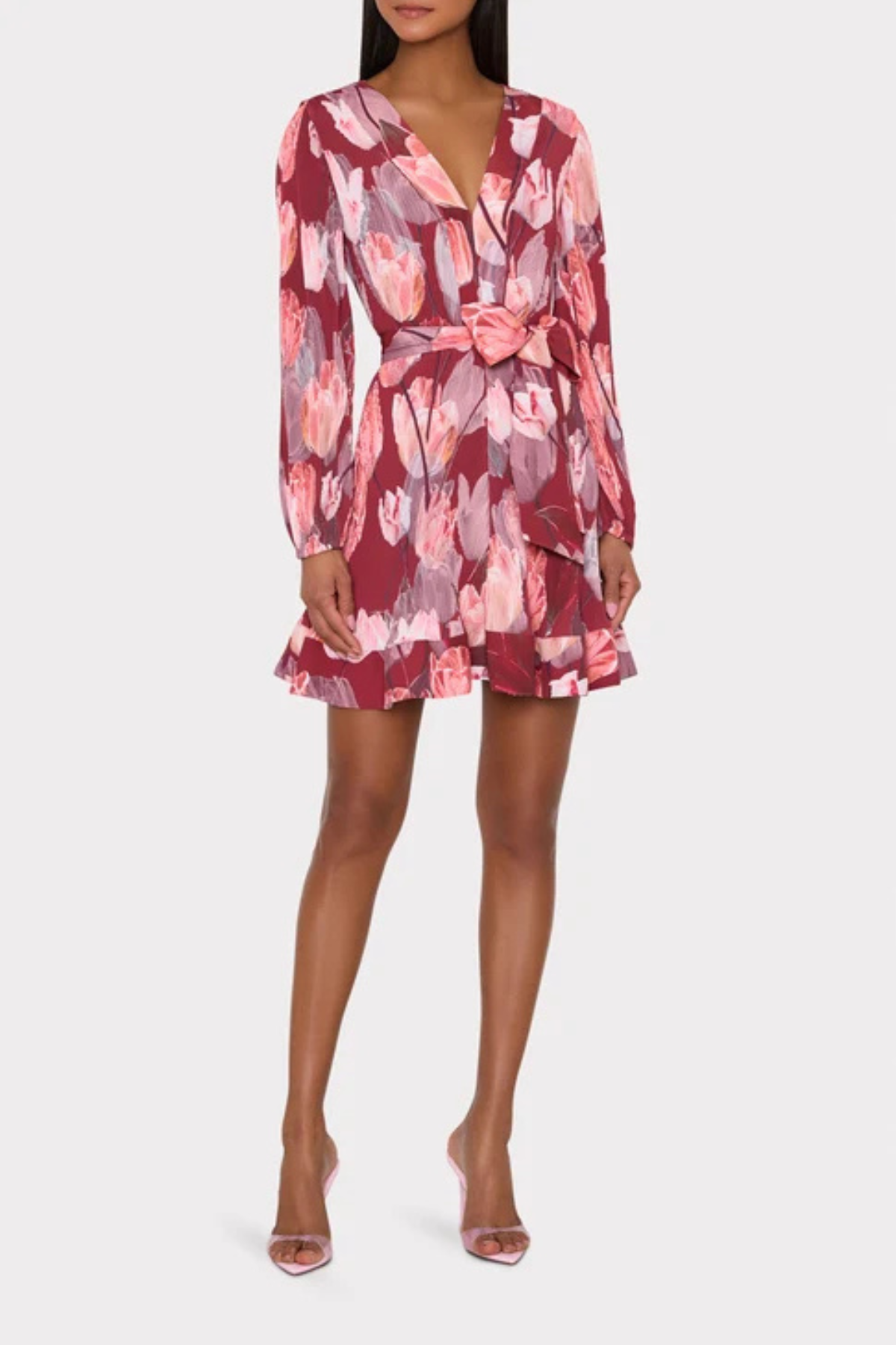 A model wearing a long-sleeve wrap-style mini dress with a pink, red, and gray floral pattern, featuring a tie waist and ruffled hem, paired with clear high-heeled sandals against a light gray background.