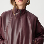 A woman wearing a shiny dark burgundy faux-leather zip-up jacket with a mock neck, photographed against a light gray studio background.