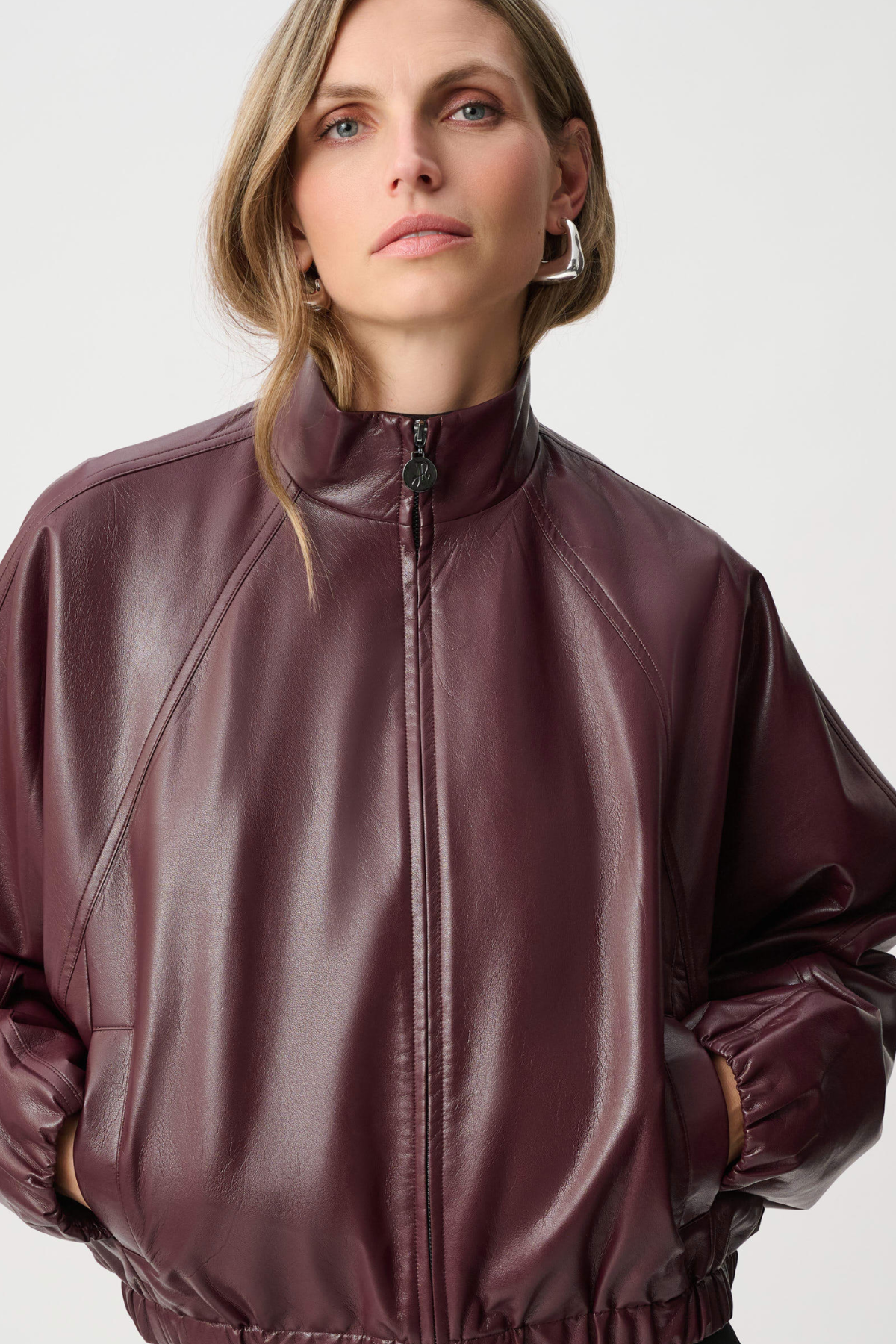 A woman wearing a shiny dark burgundy faux-leather zip-up jacket with a mock neck, photographed against a light gray studio background.