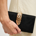 A close-up of a person holding a black clutch bag with a large, ornate gold metal clasp against a light cream-colored textured dress.