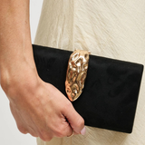 A close-up of a person holding a black clutch bag with a large, ornate gold metal clasp against a light cream-colored textured dress.