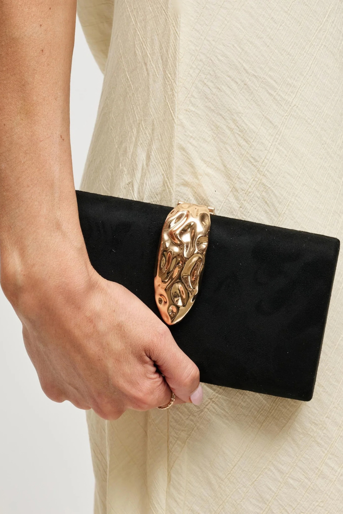 A close-up of a person holding a black clutch bag with a large, ornate gold metal clasp against a light cream-colored textured dress.