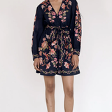 A woman modeling a navy blue long-sleeve mini dress with a pink and green floral print, standing against a plain white studio background.