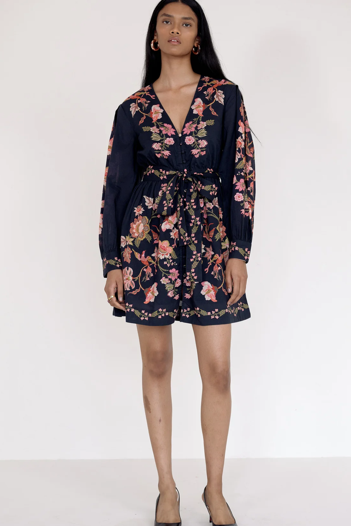A woman modeling a navy blue long-sleeve mini dress with a pink and green floral print, standing against a plain white studio background.