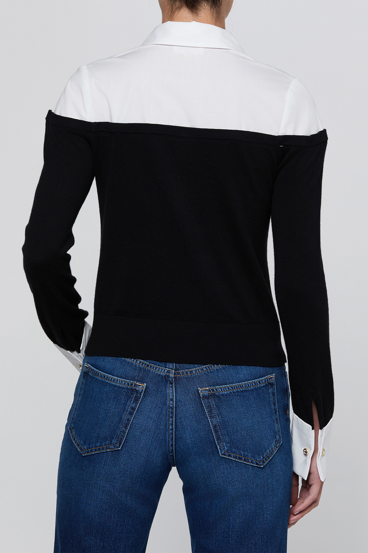 A person shown from the back wearing a color-block knit sweater with a white yoke and black sleeves, paired with blue jeans, against a light gray studio background.