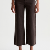 A model wearing dark brown cropped wide-leg jeans with a raw hem, styled with a white top and black loafers against a plain white studio background.