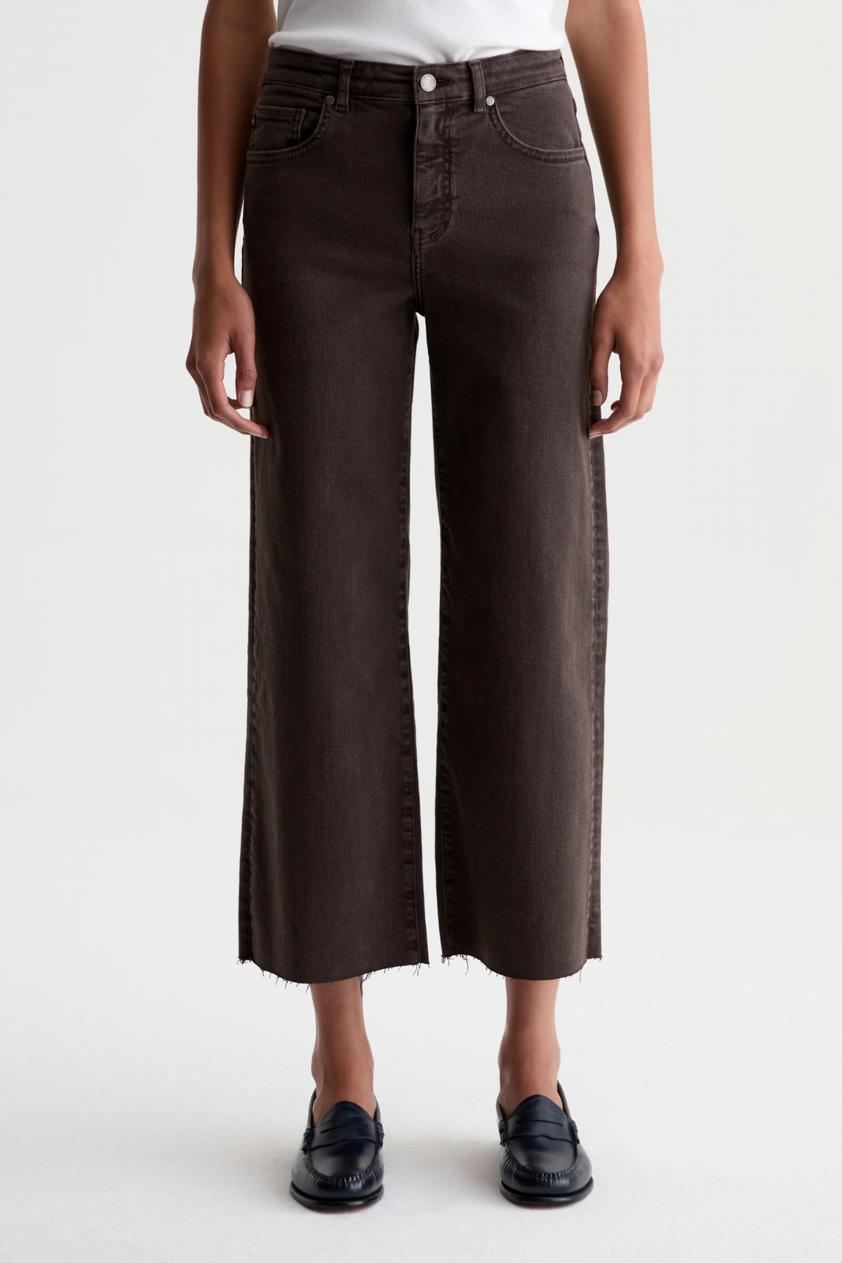 A model wearing dark brown cropped wide-leg jeans with a raw hem, styled with a white top and black loafers against a plain white studio background.