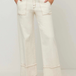 A person wearing off-white wide-leg cropped jeans with contrast stitching, paired with a navy top and black flat shoes, standing against a light gray studio background.