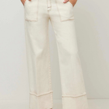 A person wearing off-white wide-leg cropped jeans with contrast stitching, paired with a navy top and black flat shoes, standing against a light gray studio background.