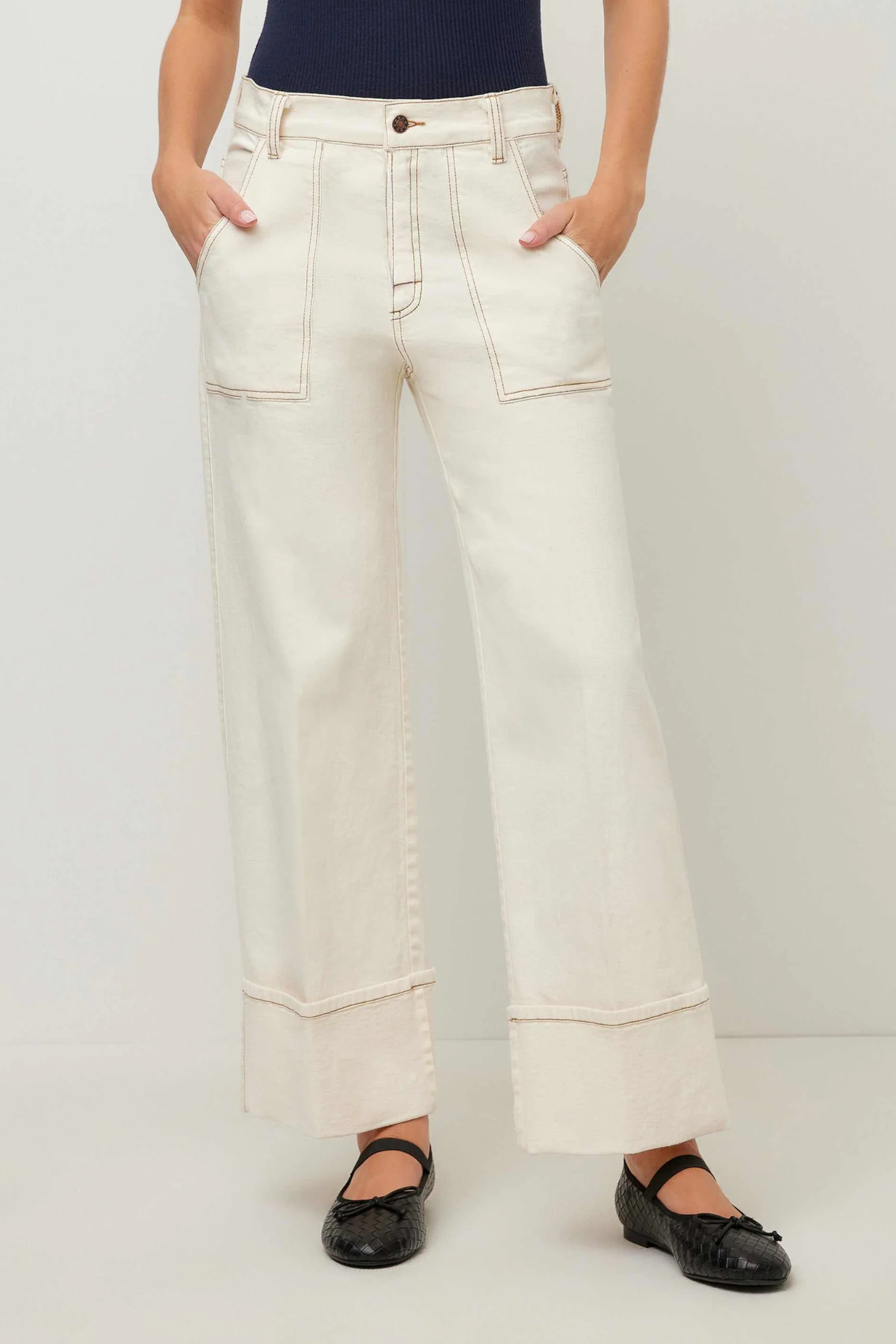 A person wearing off-white wide-leg cropped jeans with contrast stitching, paired with a navy top and black flat shoes, standing against a light gray studio background.