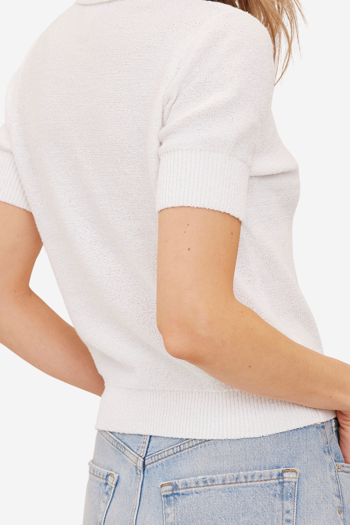 Close-up back view of a person wearing a short-sleeve white knit top with light blue jeans against a plain light background.