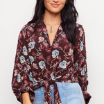 A woman modeling a dark burgundy floral tie-front blouse with blue and white flowers, paired with light-wash blue jeans, against a plain white studio background.