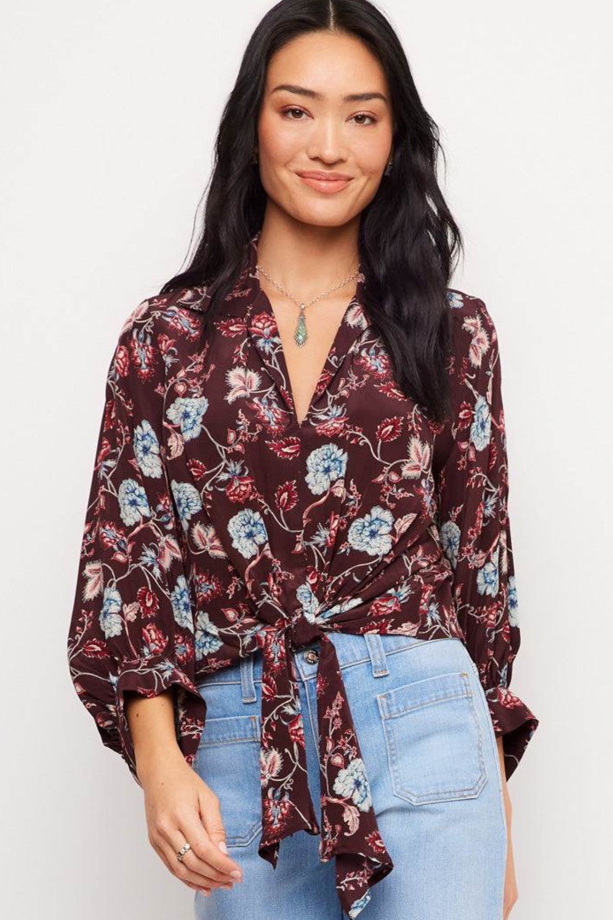 A woman modeling a dark burgundy floral tie-front blouse with blue and white flowers, paired with light-wash blue jeans, against a plain white studio background.