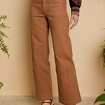 Person wearing tan pants with a patterned top and sandals, standing against a beige wall with plants.