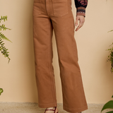 Person wearing tan pants with a patterned top and sandals, standing against a beige wall with plants.