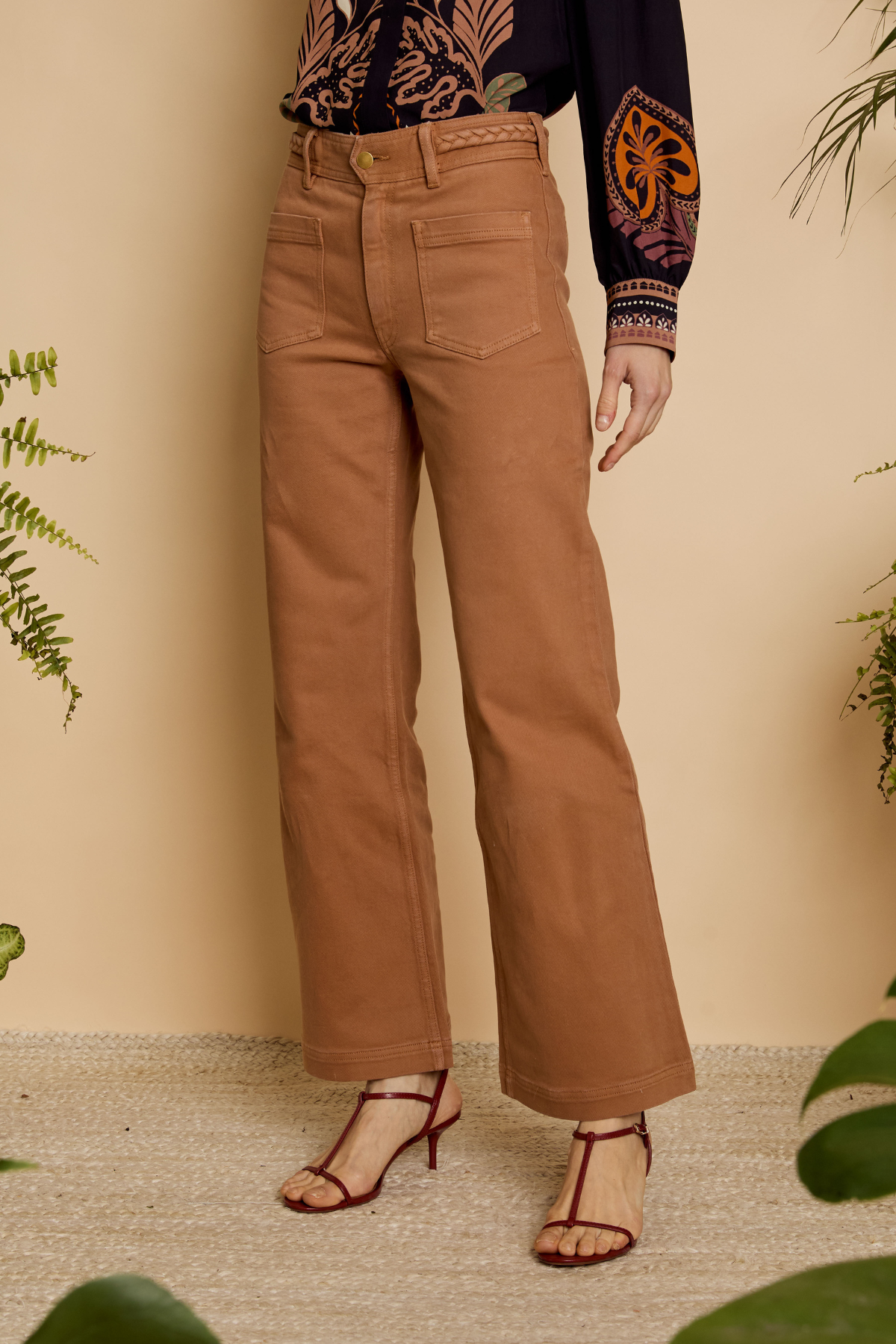 Person wearing tan pants with a patterned top and sandals, standing against a beige wall with plants.
