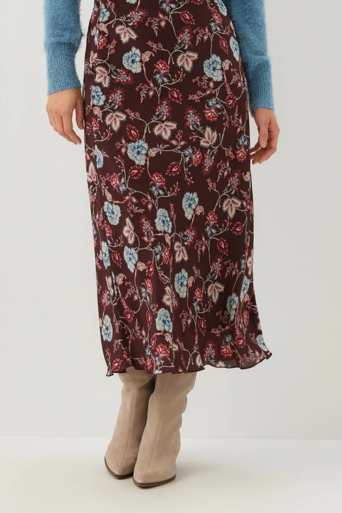 A model wearing a dark brown floral midi skirt with blue, pink, and red flowers, styled with a blue knit sweater and beige knee-high boots against a white studio background.