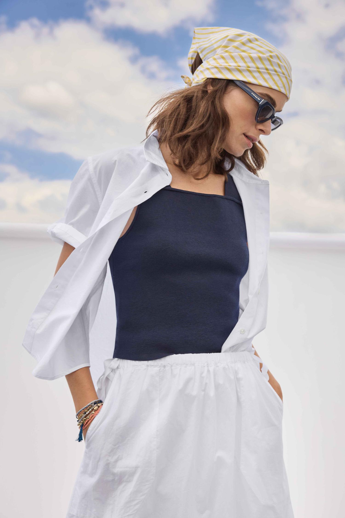 Woman wearing a white shirt, navy tank top, and white pants with a yellow and white patterned headscarf against a blue sky.