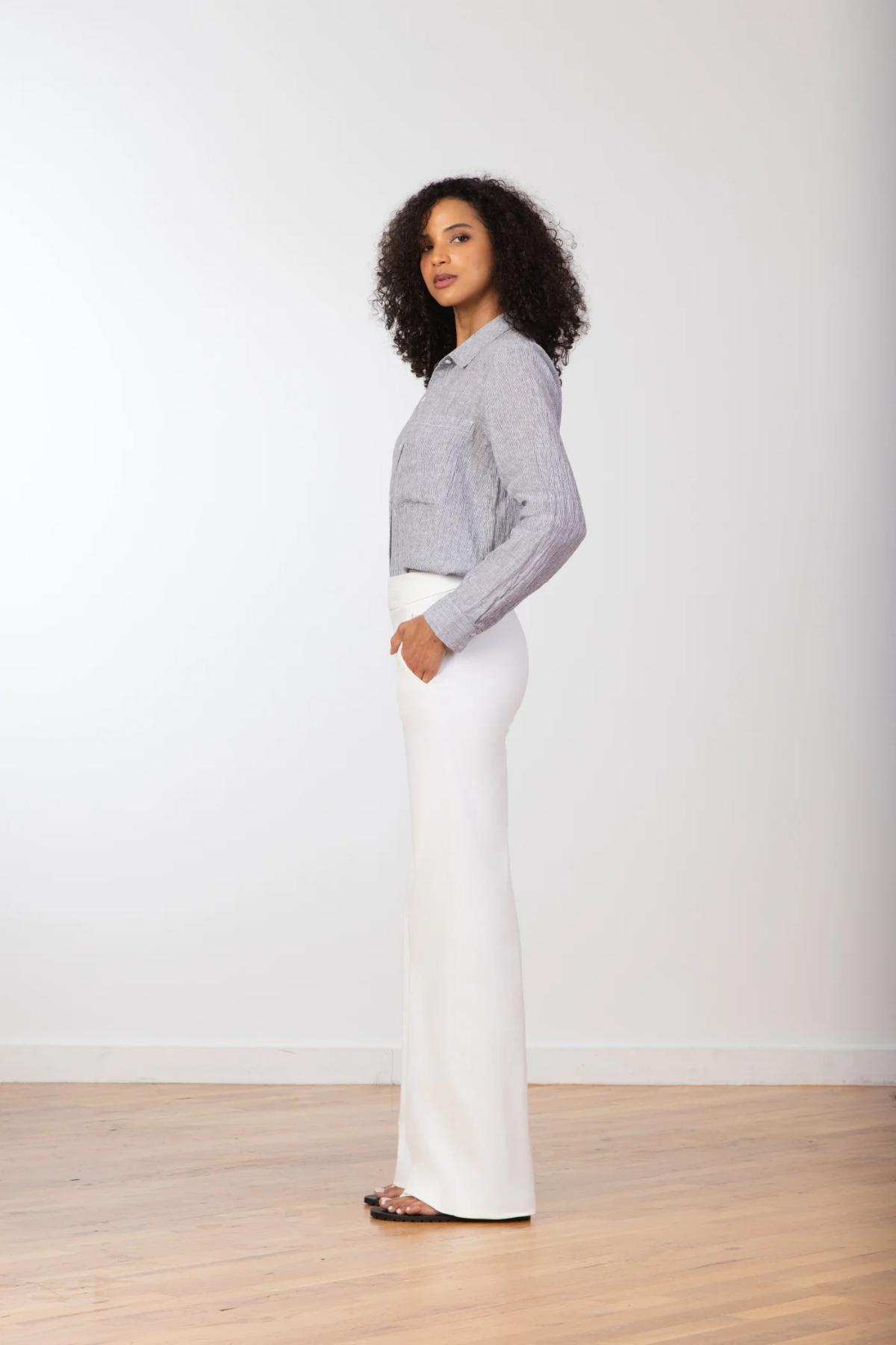 Woman wearing a gray sweater and white pants standing against a plain white background